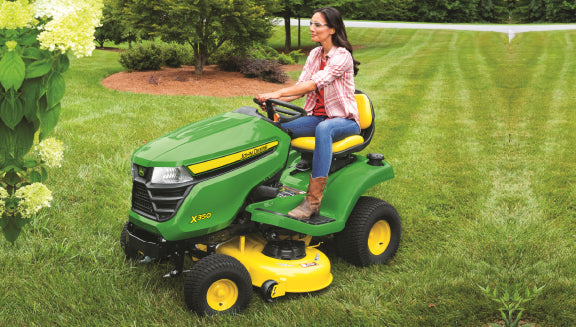 Woman wearing boots and a plaid shirt operating a green and yellow riding lawn mower on a neatly trimmed lawn with trees and bushes nearby