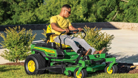 Man in a yellow shirt operating a green and yellow zero-turn lawn mower on a grassy area near shrubs and a paved surface in a residential setting