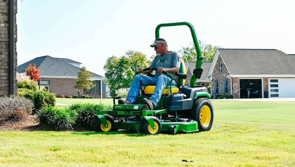 Person wearing a cap and casual clothes seated on a green and yellow zero-turn lawn mower cutting grass beside a residential driveway