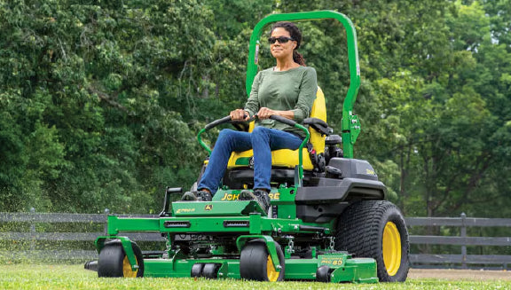 Woman wearing sunglasses and casual clothes riding a green and yellow zero-turn mower cutting grass on a spacious green lawn with trees in the background