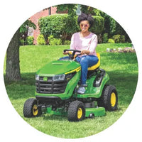 Woman wearing sunglasses and a light pink shirt sitting on a green riding lawn mower cutting grass in a sunny backyard.
