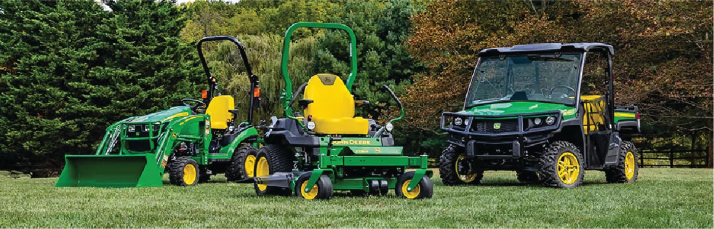 Three green and yellow outdoor machines including a compact tractor with front loader, a zero-turn mower, and a utility vehicle parked on a grassy lawn with trees in the background.