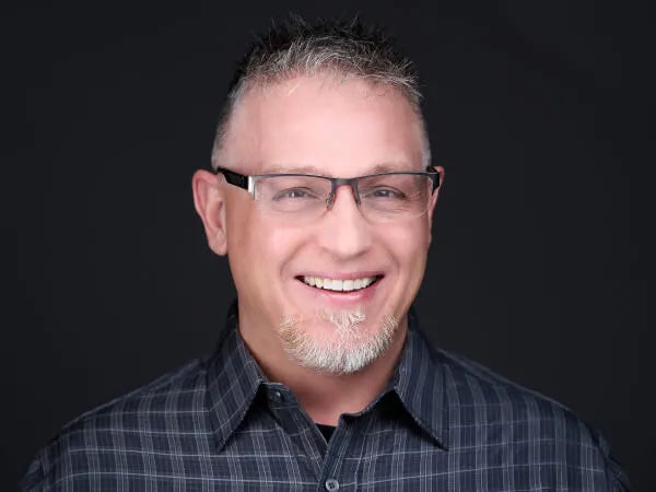 Smiling middle-aged man with short gray hair, glasses, and a dark checked button-up shirt against a plain dark background.