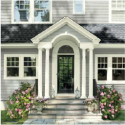 Front facade of a gray house with white trim featuring a black front door under a small portico with columns, flanked by pink flowering bushes.
