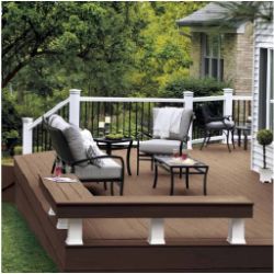 Outdoor deck with brown wooden flooring featuring cushioned metal chairs, a small glass-top table, and a serene backyard view with trees and a brick house wall.
