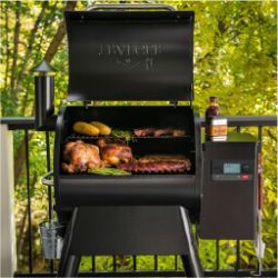 Outdoor pellet grill with open lid showing chicken, sausages, and vegetables cooking on the grates, set against a leafy backyard background.