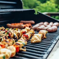 Close-up of assorted skewers with grilled vegetables and meat cooking alongside sausages on an outdoor barbecue grill with a green garden background.
