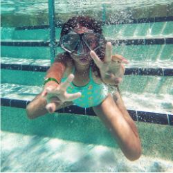 Underwater view of a child wearing a snorkel mask and blue swimsuit swimming toward the camera in a pool with steps in the background.