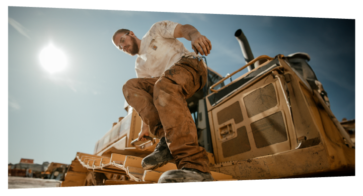 Low-angle view of a construction worker wearing dusty work clothes stepping down from heavy machinery under a bright sunlit sky, highlighting gritty outdoor work conditions.