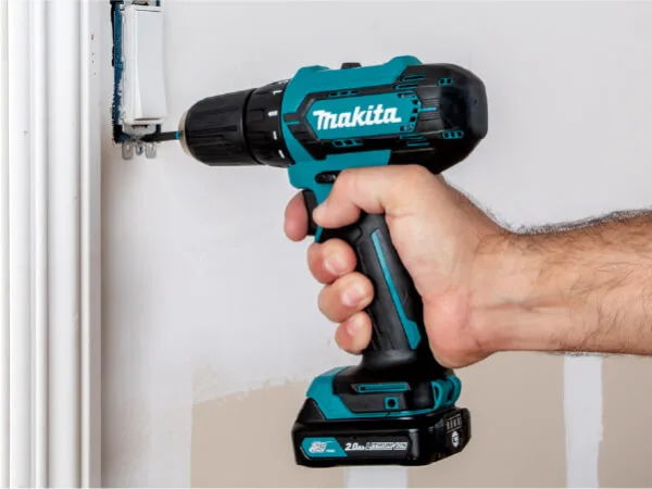 Close-up of a person using a teal and black cordless drill to install an electrical box on a white wall, demonstrating a home improvement task.