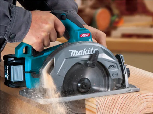 Person using a teal and gray cordless circular saw to cut a wooden beam indoors, with sawdust flying from the blade during a woodworking task.