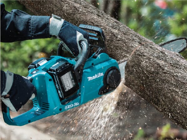 Person wearing gloves using a teal cordless chainsaw to cut a tree branch outdoors with wood chips flying during a woodworking task