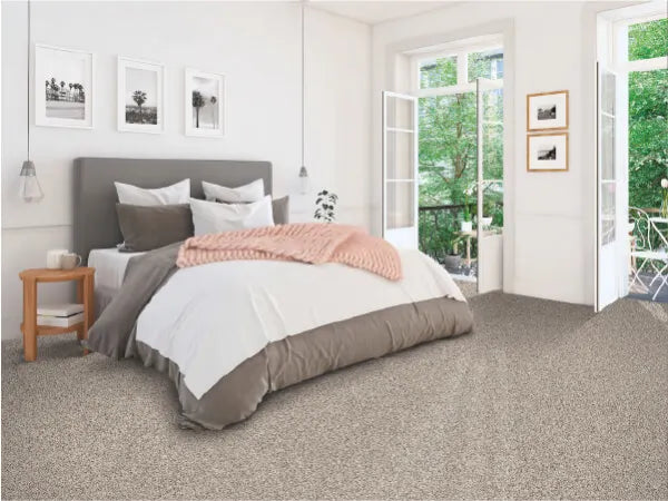 Bright, airy bedroom featuring a neatly made bed with layered bedding, beige carpet flooring, and large windows opening to a green outdoor space.