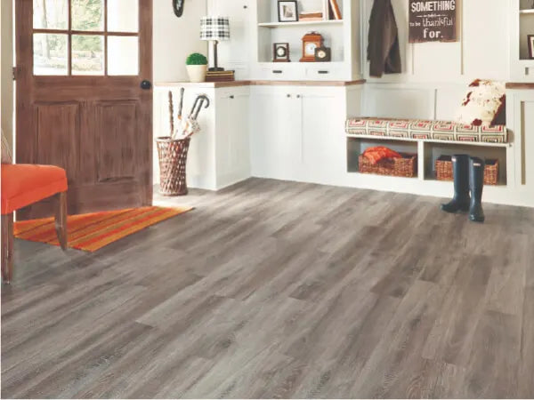 Entryway with gray wood-look flooring, a wooden door, built-in white cabinets, a patterned bench with storage baskets, and an orange chair beside a striped rug.