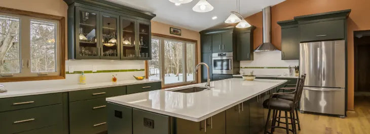 Kitchen with deep green cabinetry, large white island with integrated sink, stainless steel appliances, glass-front upper cabinets, and windows showing snowy trees outside.