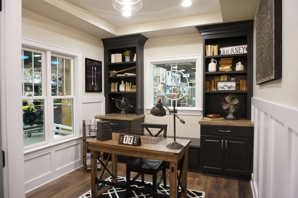 Small home office space with dark built-in shelves and cabinets flanking a window, a wooden desk with black metal legs, task lamp, decorative globe, and a geometric rug on hardwood floors.