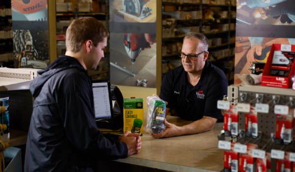 Store employee wearing a black polo shirt assisting a customer across a counter in a hardware store with tools and equipment displayed in the background.