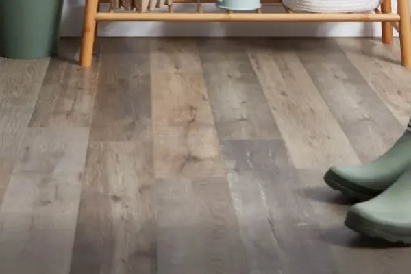 Close-up view of a wood-look vinyl flooring with varied gray and brown tones, featuring a small portion of green rubber boots and a wooden furniture piece in the background.