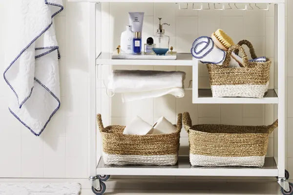 Wheeled white metal storage cart with woven baskets holding towels, brushes, and toiletries against a white paneled wall.