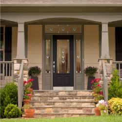 Front entrance of a house featuring a dark paneled door with decorative glass, framed by stone columns and surrounded by potted plants and greenery.