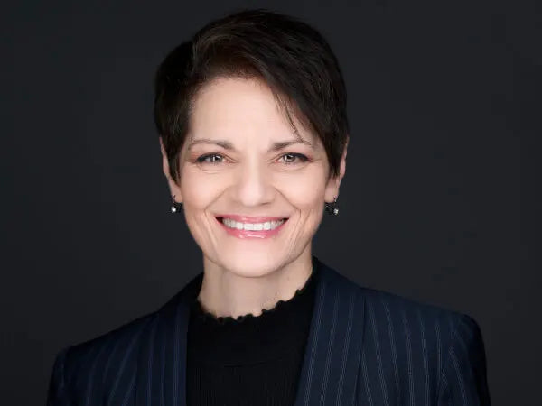 Portrait of a smiling woman with short dark hair wearing a dark pinstripe blazer and black top against a plain dark background.