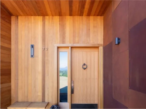 Entrance of a modern home featuring vertical wooden paneling, a natural wood door with a round door knocker, a narrow window, and the number 72 displayed next to a wall light fixture.