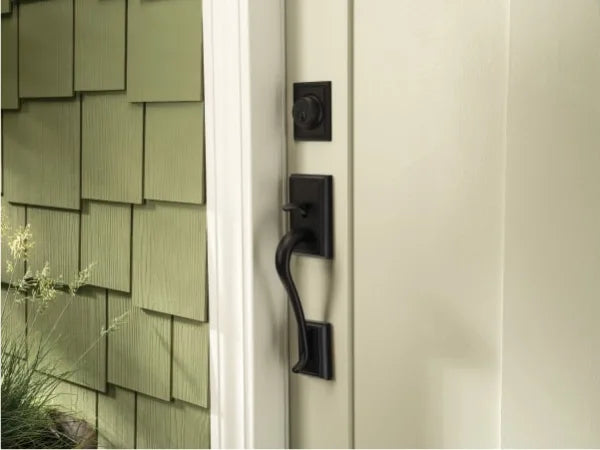 Close-up of a black metal exterior door handle and deadbolt on a light-colored door with green shingle siding visible on the side, highlighting home improvement details.