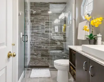 Contemporary bathroom with a glass-enclosed shower featuring dark marble tiles, white toilet, dark wood vanity with white countertop, and bright yellow flowers.