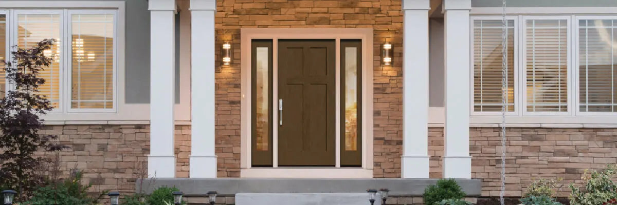 Front porch with a brown door featuring narrow sidelights, flanked by white columns and warm exterior lighting on a stone and siding facade.