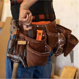 Close-up of a brown leather tool belt worn around the waist, holding various hand tools including pliers, a hammer, a level, a ruler, and safety glasses during a home improvement project.