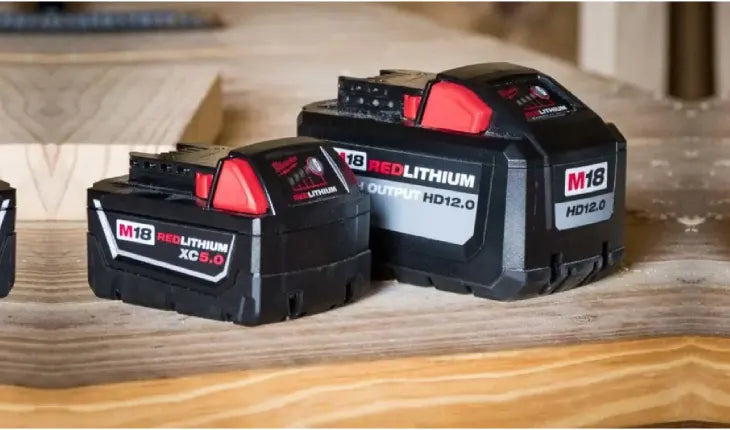 Two black and red rechargeable lithium-ion batteries labeled M18 Red Lithium placed on a wooden surface in a workshop setting.