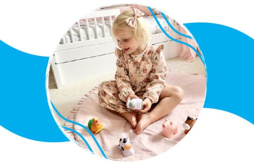 Young child sitting on a pink blanket in a bright room, playing with small colorful animal toys near a white crib.