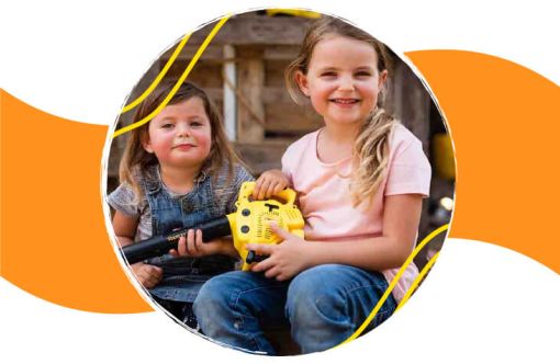 Two young children sitting outdoors, smiling and playing together with a yellow toy resembling a handheld power tool.