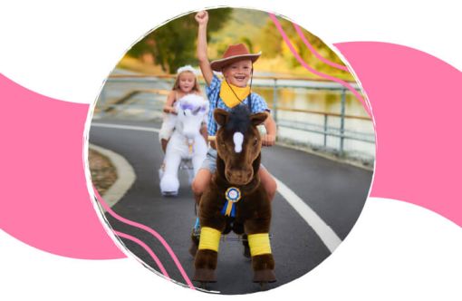 Two children dressed in costumes riding motorized toy animals along a paved outdoor path with a metal railing.