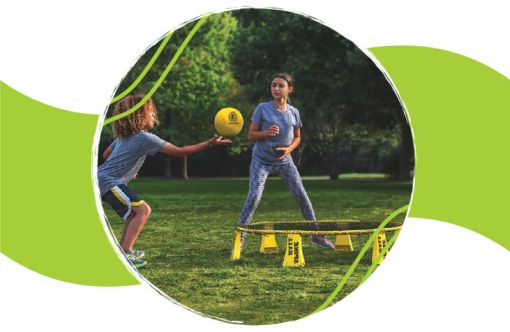 Two children playing a game of spikeball on grass outdoors with trees in the background, one preparing to hit a yellow ball.