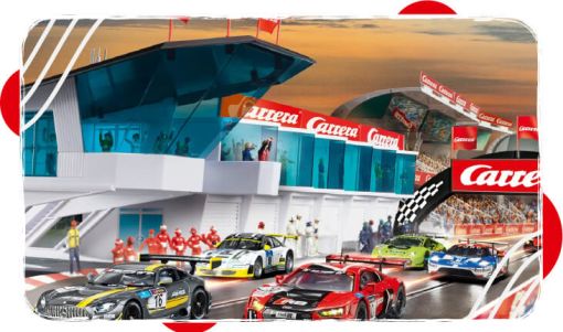 Race cars lined up on a track in front of a modern glass-paneled control tower with spectators and team members in the background.