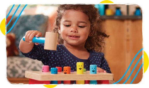 Young child smiling while playing with a wooden hammer toy featuring colorful, smiling pegs in a cozy indoor setting.
