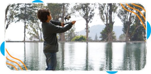 Young boy casting a fishing rod while standing near the edge of a calm lake surrounded by trees and distant hills.
