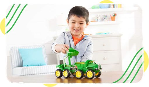 Young boy playing indoors with a green toy tractor and trailer on a wooden table in a bright living room setting
