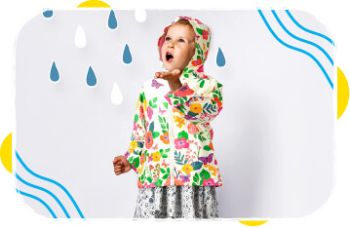 Young girl wearing a colorful floral raincoat with a hood, standing indoors against a white wall decorated with blue and white raindrop shapes.