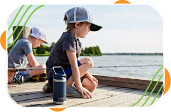 Two young boys wearing caps sitting on a wooden dock by the water, one holding a dark insulated bottle beside him on a sunny day.