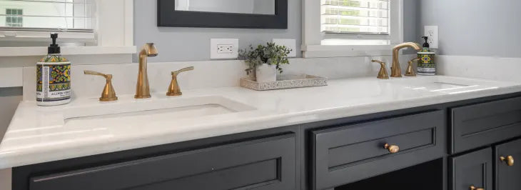 Double vanity with a white countertop, brass faucets, dark gray cabinetry, decorative soap dispensers, and two windows above with white blinds.