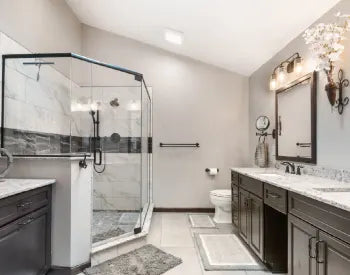 Modern bathroom featuring a frameless glass shower with tile accents, dual dark wood vanities with marble countertops, and muted gray walls.