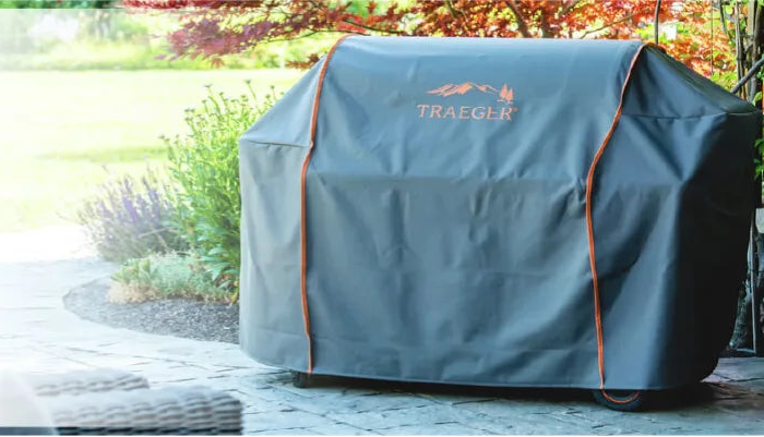 Outdoor scene showing a covered grill with a dark protective cover featuring orange trim and a logo, set on a stone patio near greenery.