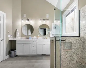Bright transitional bathroom with white cabinetry, double round mirrors, marble countertop, wall-mounted lights, and glass shower enclosure with granite tiles.