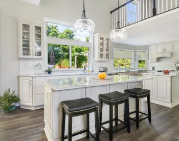 Light-filled kitchen featuring white cabinetry, large central island with quartz countertop, black leather stools, pendant lighting, and wide windows overlooking greenery.