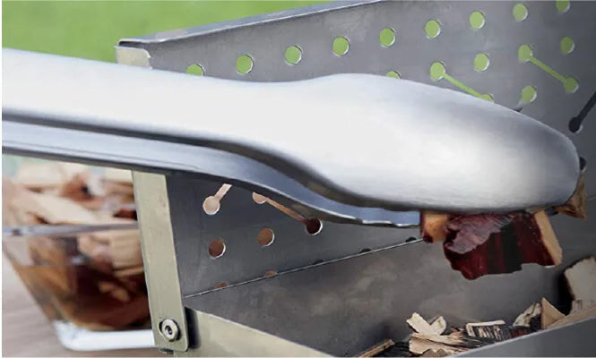 Close-up of a stainless steel grilling tool resting on the edge of a metal smoker box filled with wood chips, outdoors on grass.