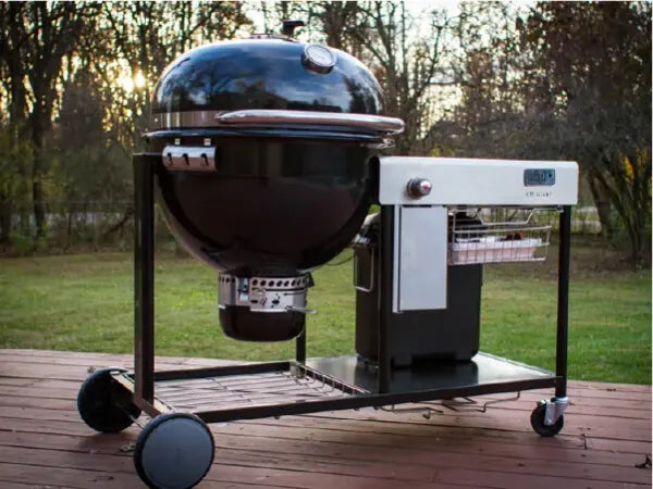Black round charcoal grill with an attached rectangular side compartment on a wheeled metal frame, set outdoors on a wooden deck with trees in the background.