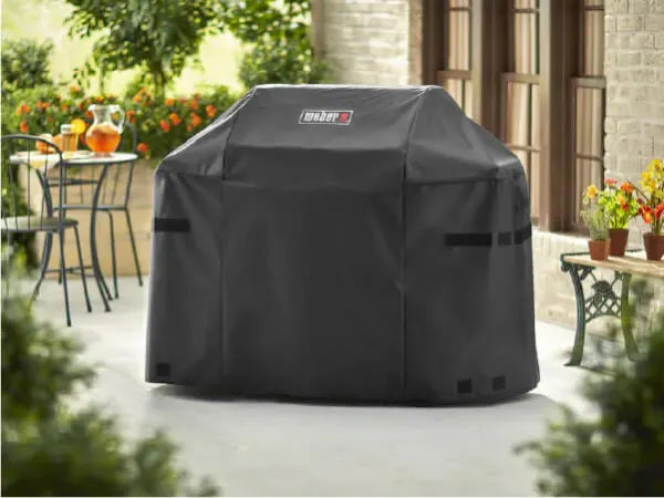 Black weather-resistant grill cover with a logo on the front, protecting a grill on a patio surrounded by plants and outdoor furniture.