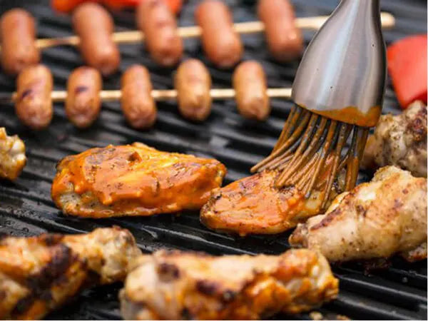 Grilled chicken pieces being brushed with sauce using a basting brush on a barbecue grill with sausages cooking in the background.